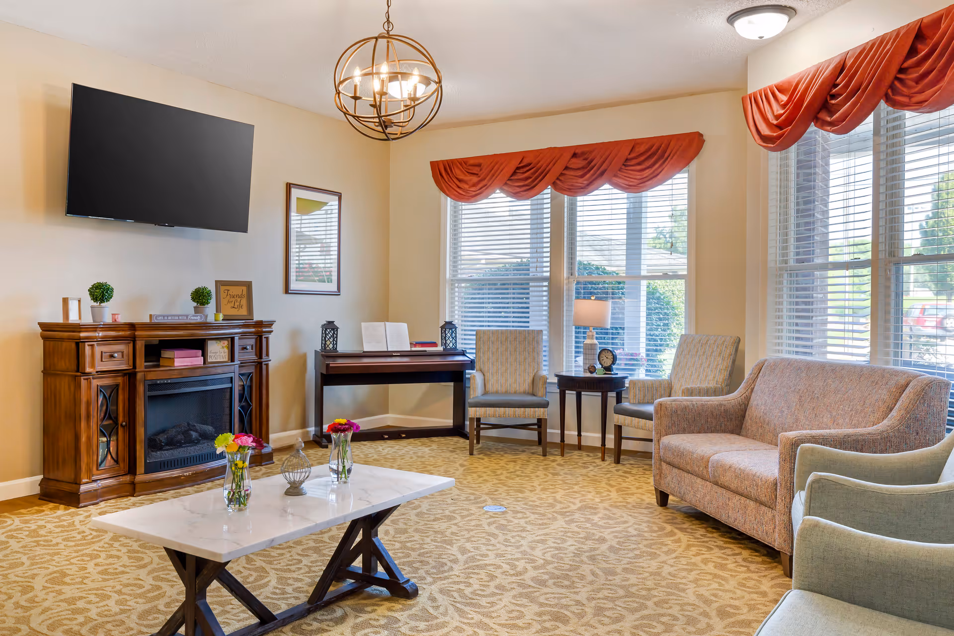 A bright senior living lounge with sofas and armchairs around a coffee table, a wall-mounted TV above a wooden fireplace cabinet, and large windows with red valances.