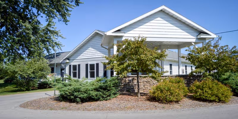 Exterior view of a single-story building with white siding and a covered entrance, surrounded by green bushes and trees under a clear blue sky.
