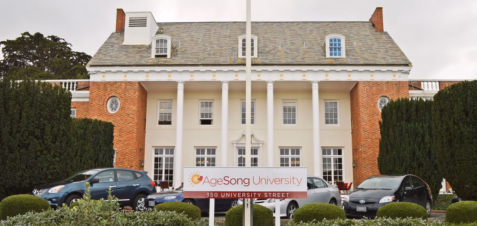 Front exterior view of a large, stately brick building with white columns and multiple windows. Several cars are parked in front, and there is a sign that reads 'AgeSong University 350 University Street'.