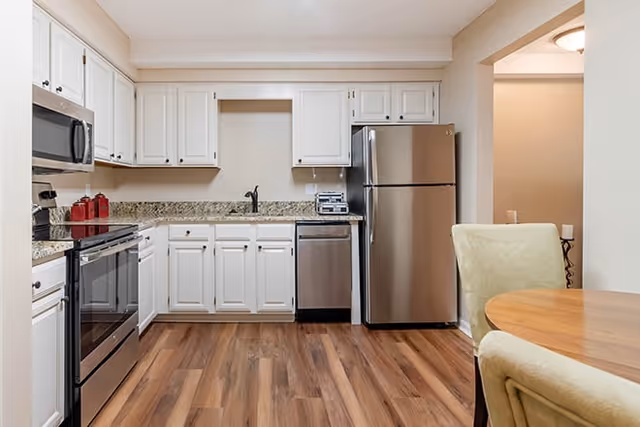 A modern kitchen with white cabinets, granite countertops, stainless steel appliances including a refrigerator, dishwasher, microwave, and stove. The floor is wood laminate, and there is a round wooden dining table with two upholstered chairs partially visible on the right.