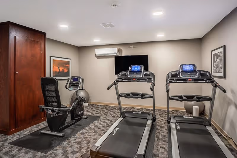 Indoor fitness room with two treadmills and one recumbent exercise bike on carpeted floor. A wall-mounted TV and an air conditioning unit are visible on the wall. The room has neutral-colored walls with framed pictures and a wooden door on the left side.