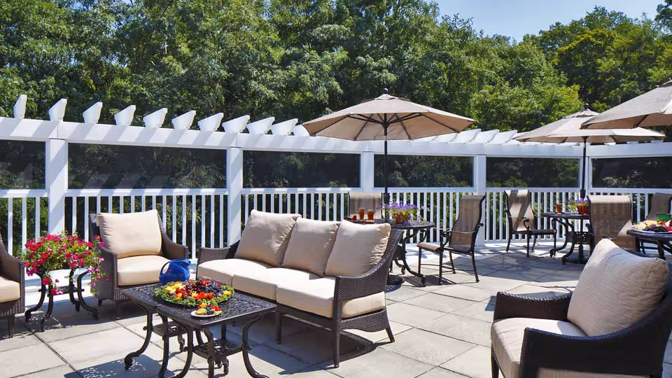 Outdoor patio area with cushioned wicker chairs and sofas arranged around black metal tables. Some tables have umbrellas providing shade. There are trees in the background and a white railing enclosing the patio.