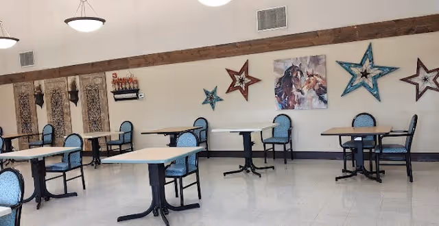 A dining room with several tables and blue cushioned chairs arranged neatly. The walls are decorated with large star-shaped ornaments and a painting of horses. The room has a high ceiling with hanging light fixtures and wooden beams along the top of the walls.