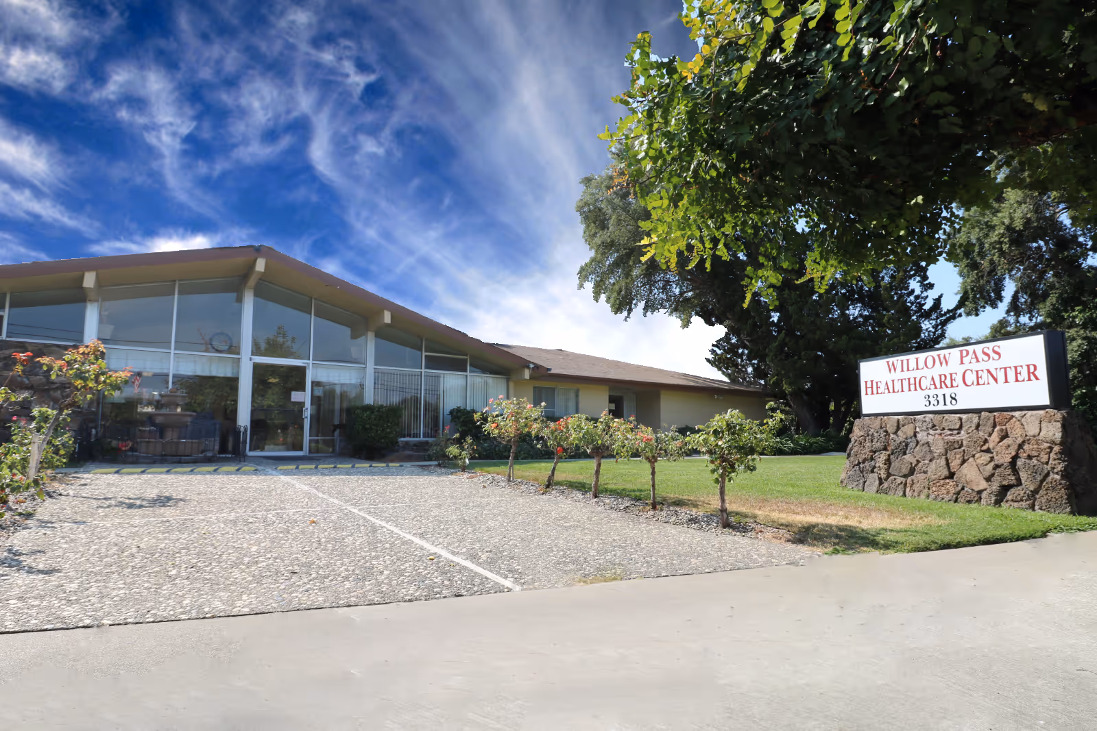 Exterior view of Willow Pass Healthcare Center showing a single-story building with large glass windows and a stone sign with the facility's name and address. The foreground includes a paved driveway and landscaped greenery with small bushes and a large tree.