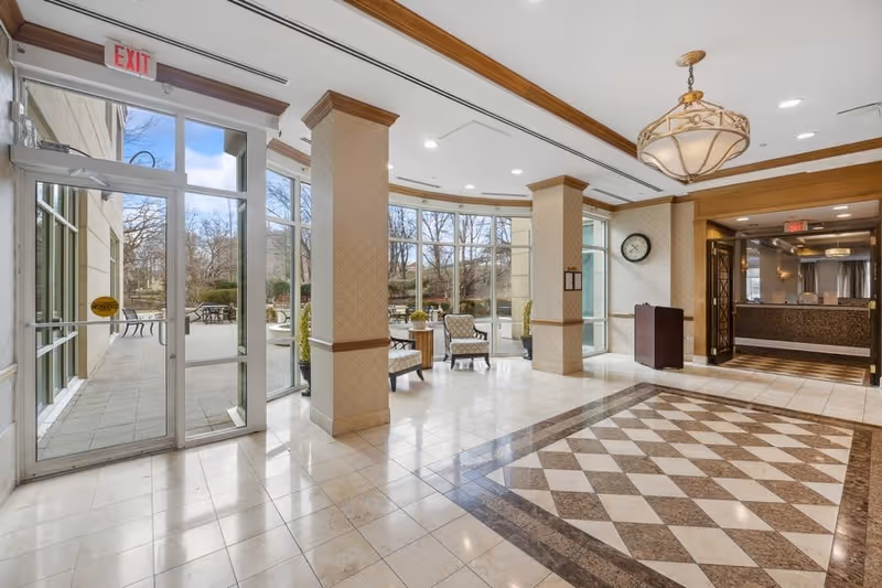 Bright and spacious lobby area with large windows showing an outdoor patio with tables and chairs. The interior features tiled floors with a diamond pattern, beige walls with wooden trim, a hanging light fixture, and a clock on the wall. There is a reception desk visible in the background and an exit door leading outside.