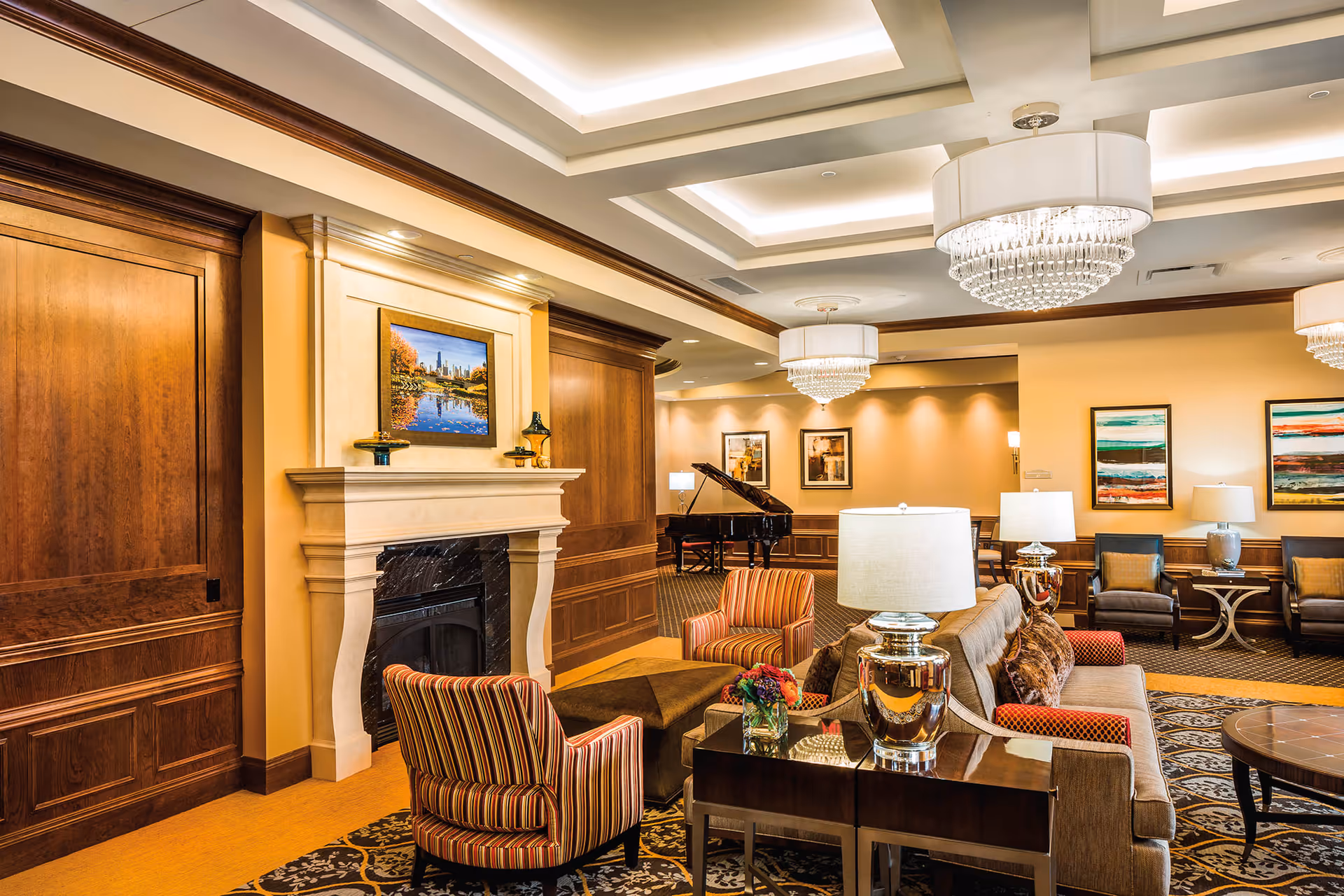 A spacious and elegant living room area featuring a fireplace with a framed picture above it, comfortable seating including striped armchairs and a sofa, decorative lamps on side tables, a grand piano in the background, and modern chandeliers hanging from a coffered ceiling.