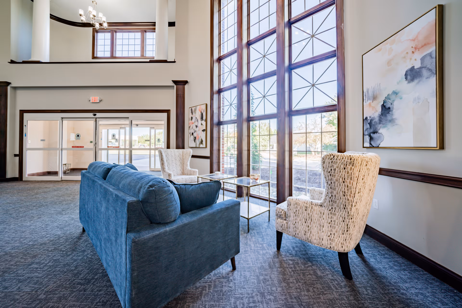 A bright and spacious seating area in a senior living facility with large floor-to-ceiling windows letting in natural light. The area features a blue upholstered sofa, two patterned armchairs, and a glass coffee table. There are decorative paintings on the walls and an exit door in the background.
