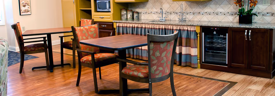 Interior view of a dining area in a senior living facility with wooden tables and chairs featuring red and green leaf-patterned upholstery. The room has a wooden floor, a kitchenette with a sink, microwave, and wine cooler, and a decorative curtain under the countertop. A framed picture is visible on the wall.