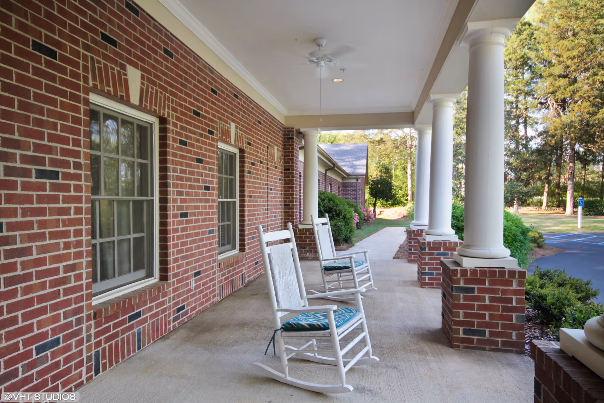 Covered brick-front porch with white columns and rocking chairs overlooking landscaped grounds.