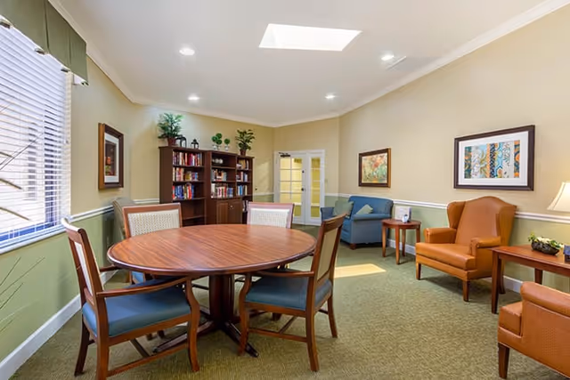 A cozy common area with a round wooden table surrounded by four chairs with blue cushions. In the background, there is a wooden bookshelf filled with books and decorative plants on top. To the right, there are two armchairs, one blue and one orange, with side tables and framed artwork on the walls. The room has beige walls with white trim, a green carpet, and a skylight in the ceiling providing natural light.