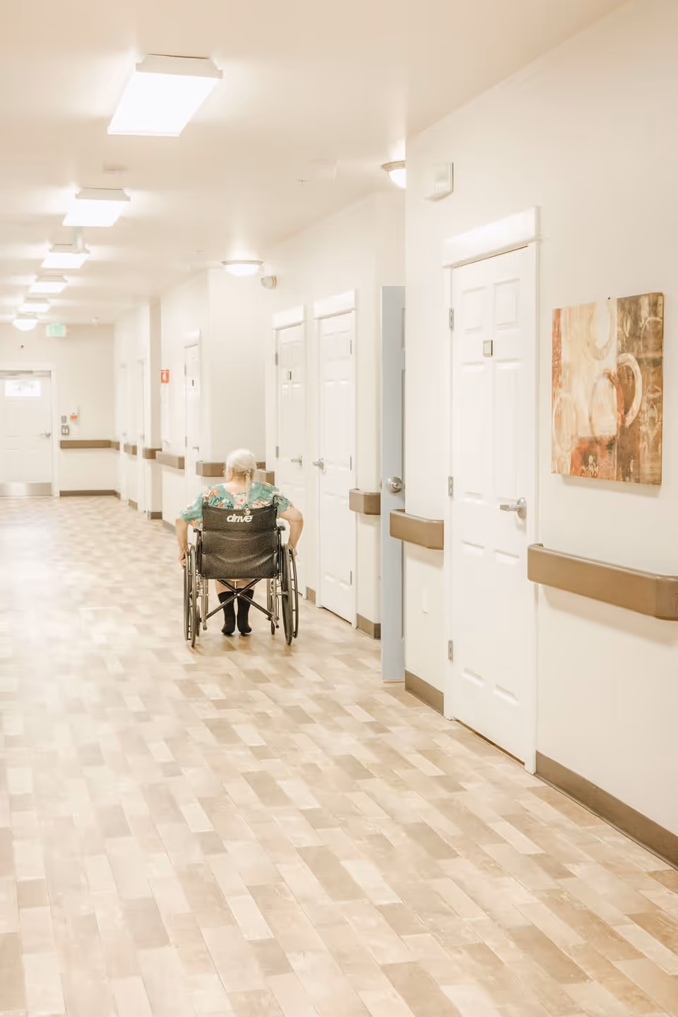 A long, well-lit hallway in a senior living facility with light-colored walls and wooden flooring. An elderly person with white hair, wearing a floral shirt, is seen from behind sitting in a wheelchair moving down the hallway. Several closed white doors line the hallway, and a piece of abstract artwork is mounted on the wall to the right.