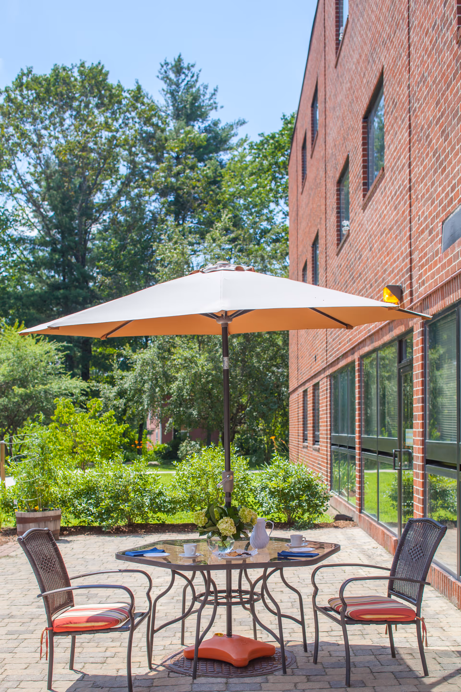 Patio dining table with umbrella and two chairs on a paved courtyard beside a brick building and green landscaping.