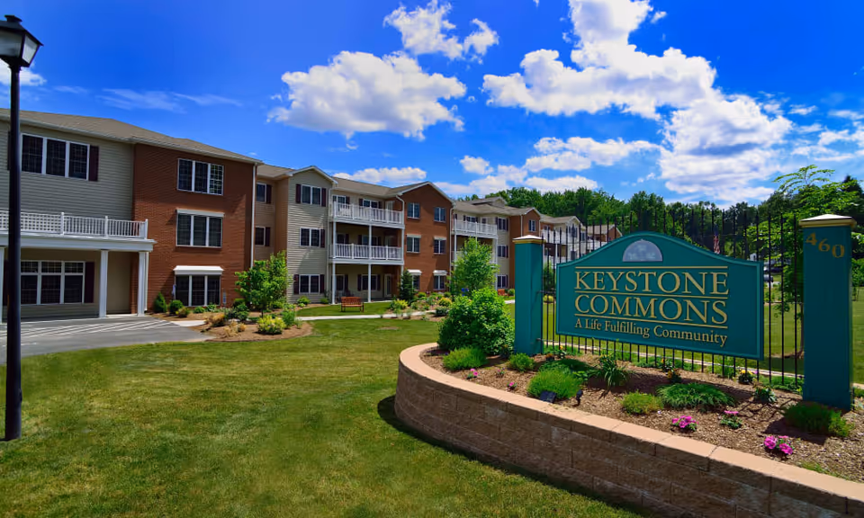 Exterior view of Keystone Commons senior living facility on a sunny day with a blue sky and scattered clouds. The building is three stories tall with a combination of brick and beige siding, balconies, and large windows. In the foreground, there is a landscaped area with green grass, shrubs, flowers, and a green sign that reads 'Keystone Commons A Life Fulfilling Community'.