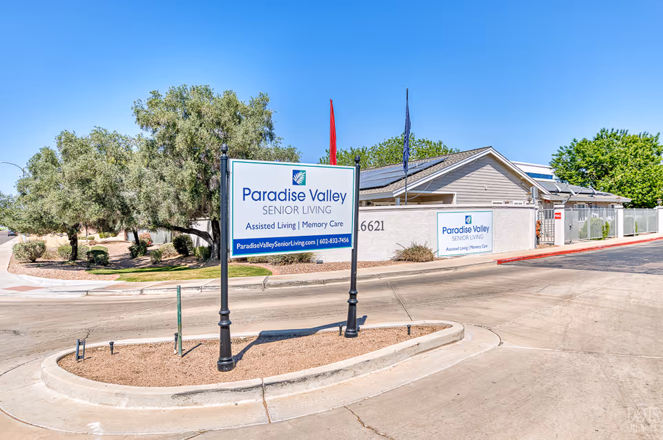 Exterior view of Paradise Valley Senior Living facility with a large sign displaying the name, services offered including Assisted Living and Memory Care, and contact information. The building is single-story with solar panels on the roof, surrounded by trees and a clear blue sky.