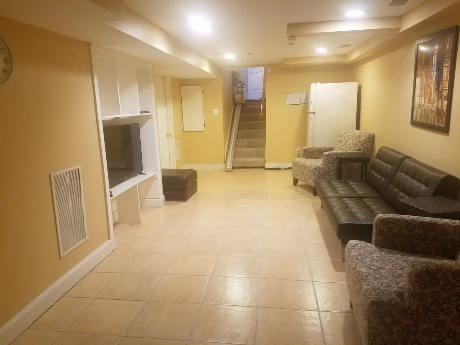 Basement living area with tiled floor, a TV in a built-in shelf, a leather sofa and patterned armchairs, and stairs at the back.