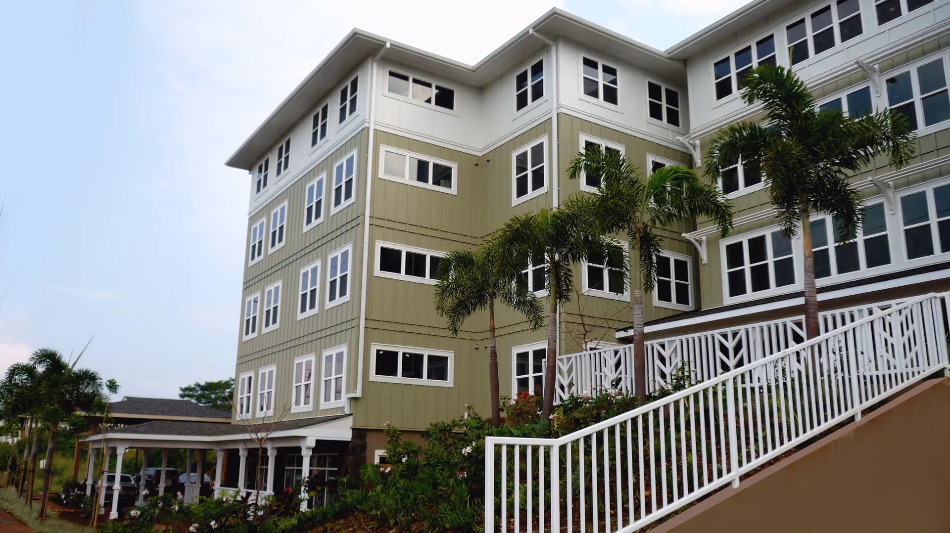 Exterior view of a multi-story senior living facility building with green siding and numerous white-framed windows, surrounded by palm trees and landscaped greenery, with a white railing along a sloped walkway.