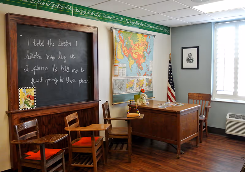 A vintage classroom setup with wooden desks and chairs, a large blackboard with cursive writing, a world map hanging on the wall, an American flag in the corner, and a wooden teacher's desk with a stuffed animal and books on it. The room has wood flooring and a window with blinds.