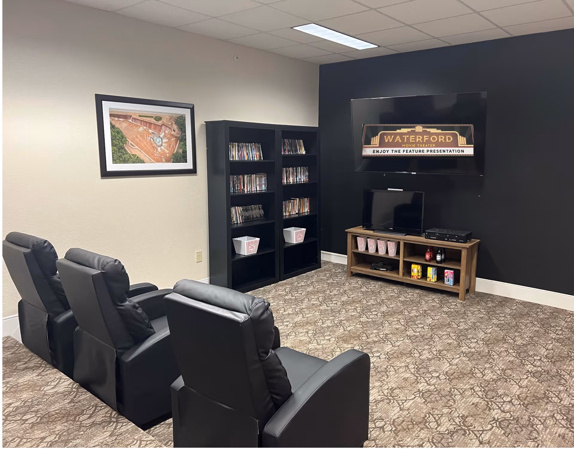 A small movie theater room with three black recliner chairs facing a wall-mounted flat screen TV. Below the TV is a wooden media console holding a smaller TV, popcorn containers, and various snacks. Two black bookshelves filled with DVDs and popcorn containers are against the adjacent wall. The room has patterned carpet and a framed aerial photo on the wall.