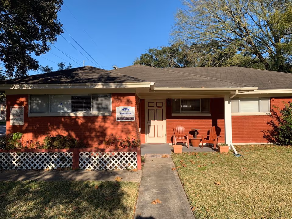 Single-story red brick building with a gray shingled roof, a small front porch with two red chairs and a table, a concrete walkway leading to the front door, and a grassy lawn with some plants and trees around.