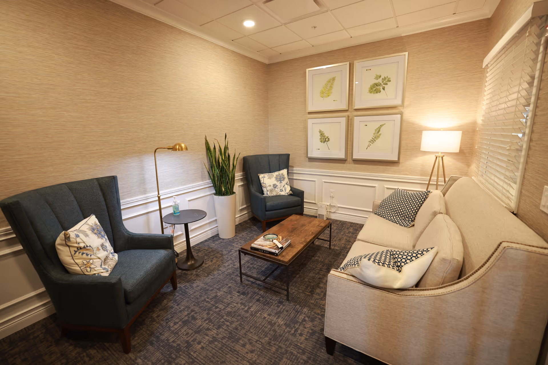 A cozy sitting area with a beige sofa and two dark blue armchairs, each with decorative pillows. A wooden coffee table with books is in the center on a patterned carpet. The walls are light beige with white wainscoting, decorated with four framed botanical prints. A tall floor lamp and a potted plant add warmth to the space.