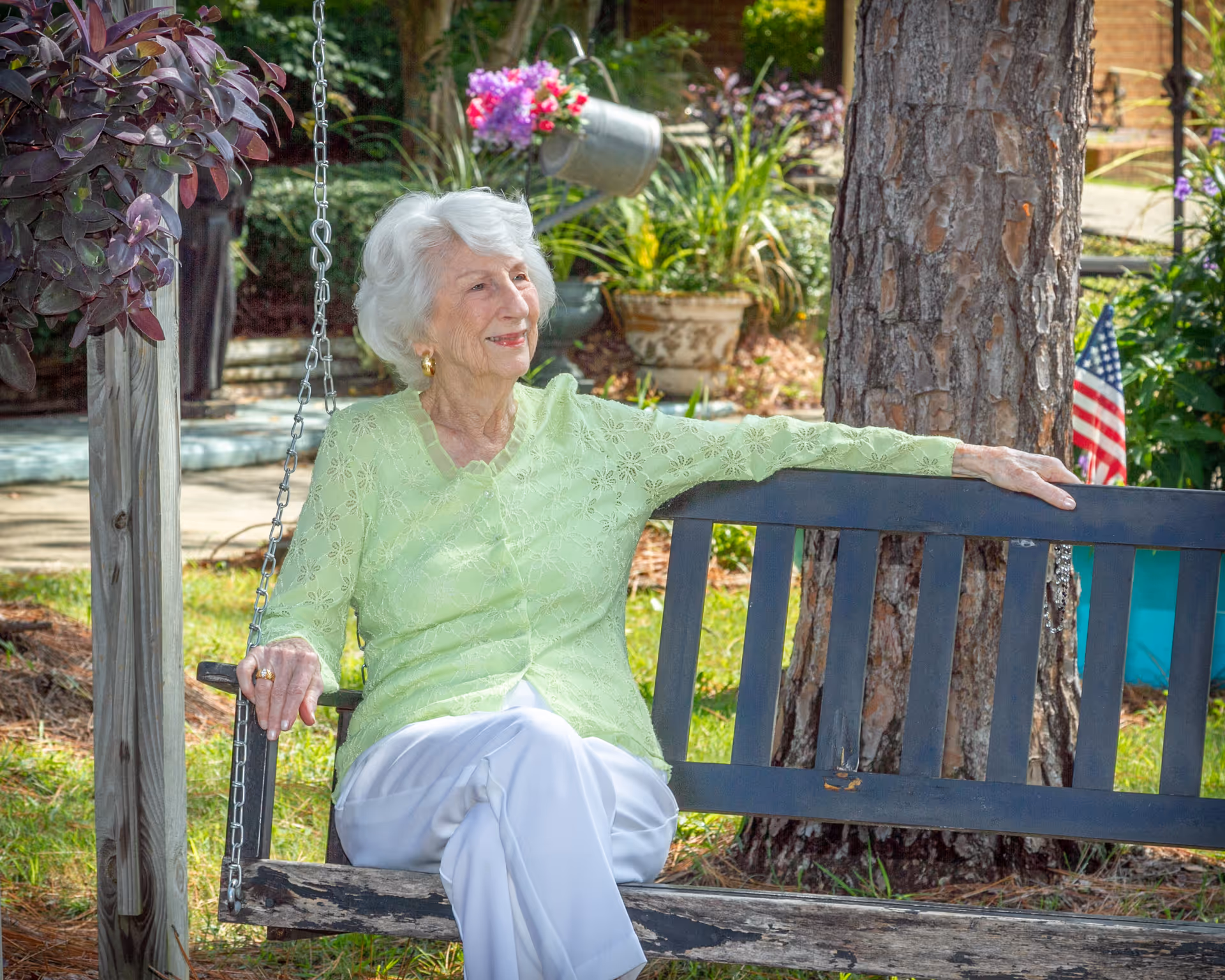 An elderly woman with white hair wearing a light green blouse and white pants is sitting on a wooden swing bench in a garden. She is smiling and looking to her left. The background includes a tree, potted plants, and an American flag.