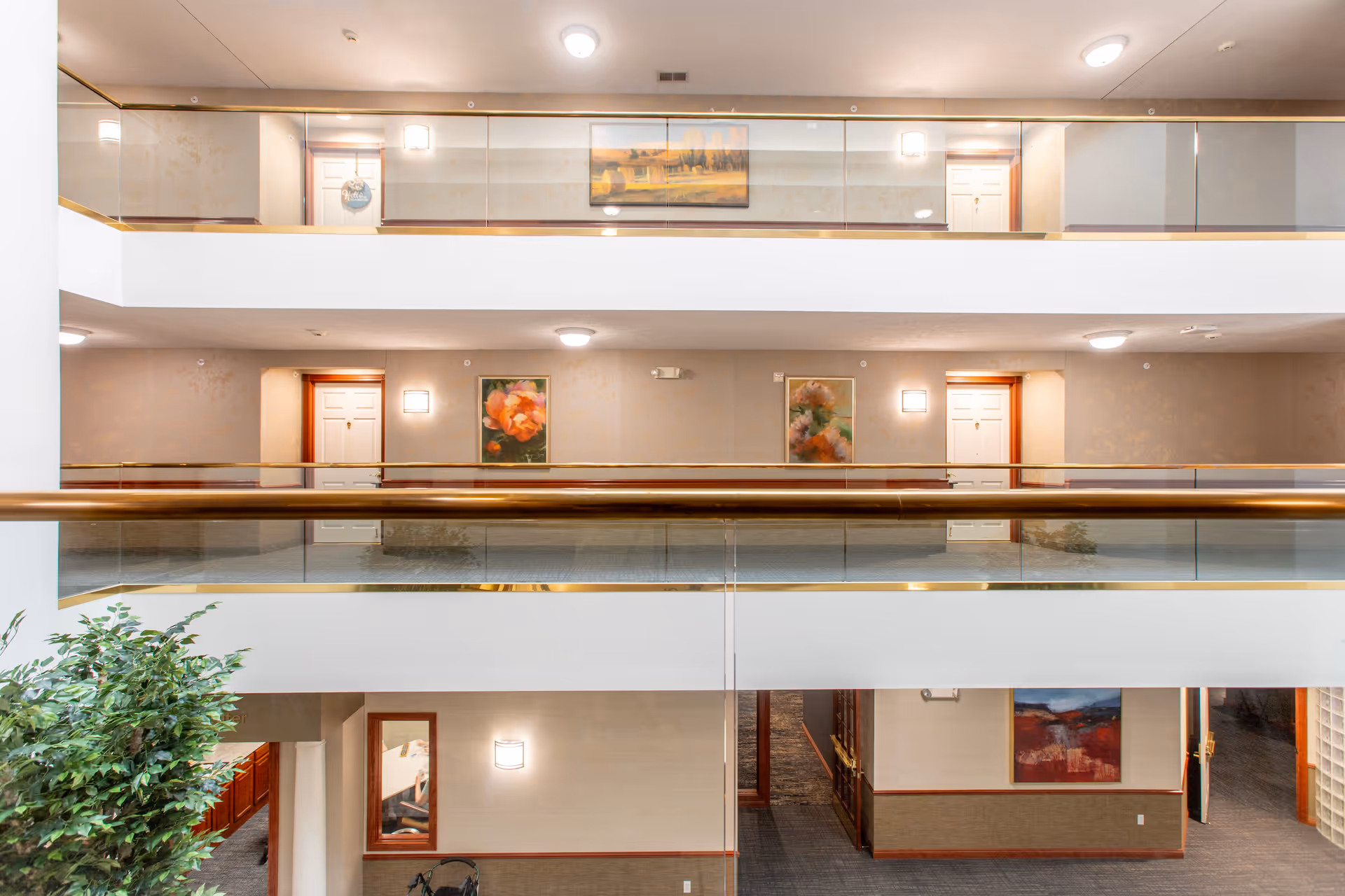 Interior view of a multi-level hallway in a retirement community with white walls, glass and brass railings, several closed doors, framed floral artwork on the walls, ceiling lights, and a large indoor plant on the lower level.