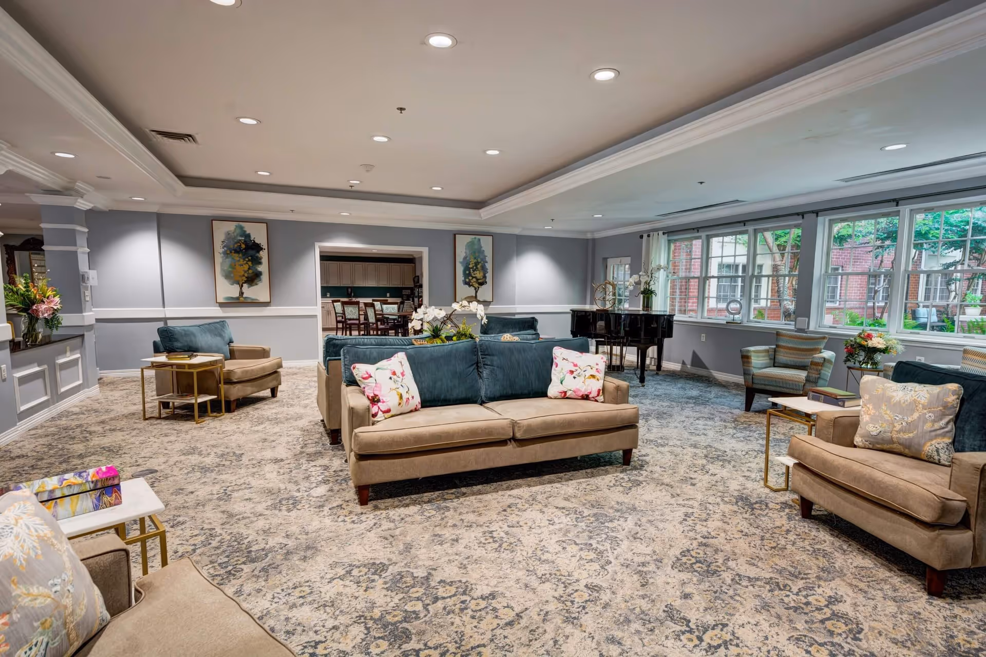 A spacious and well-lit living room area in Magnolia Place of Roswell featuring multiple beige and teal sofas and armchairs with floral and patterned cushions, a patterned carpet, large windows letting in natural light, a black grand piano near the windows, and decorative paintings on the walls.
