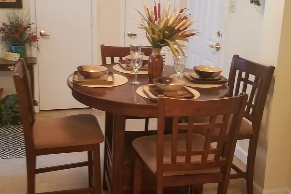 A dining area with a round wooden table set for four people. Each place setting includes a bowl, plate, and glass on a placemat. A vase with decorative flowers is in the center of the table. Four wooden chairs with cushioned seats surround the table. The background shows a white door and part of a small side table with a plant and decorative items.