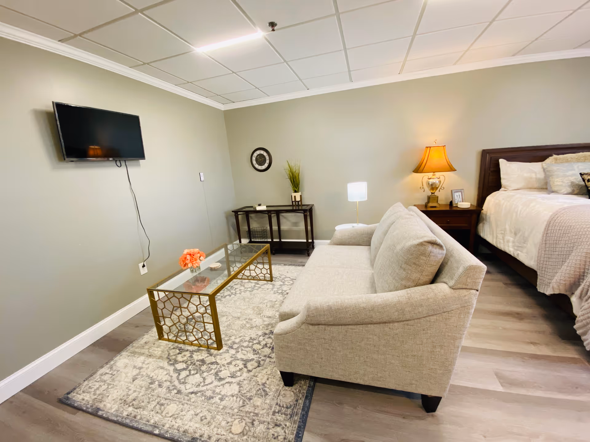 A cozy senior living room and bedroom area featuring a beige sofa facing a wall-mounted flat-screen TV. There is a glass coffee table with a decorative flower arrangement on a patterned rug. In the background, a bed with white and beige bedding is visible next to a nightstand with a lamp and framed photo. The walls are light gray, and the floor is wood laminate.