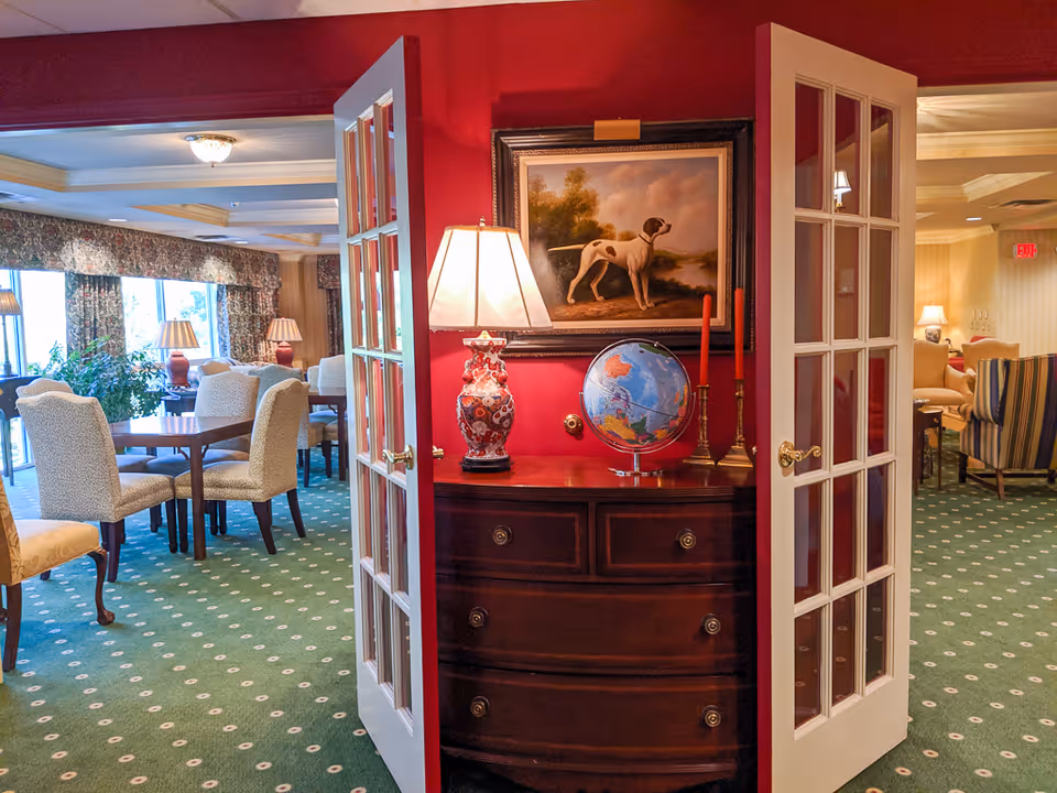 Interior view of a senior living community common area with an open double glass door. Inside, there is a wooden chest of drawers with a decorative lamp, a globe, and two red candles on top. Above the chest hangs a framed painting of a dog. The room beyond has green carpet with a white pattern, several upholstered chairs, tables, and large windows with floral curtains letting in natural light.