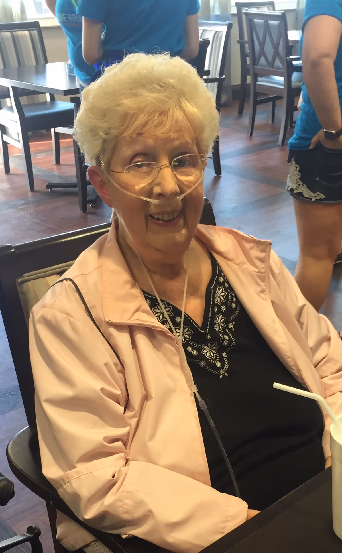 An elderly woman wearing a nasal oxygen cannula sits and smiles at a dining table in a communal dining area.