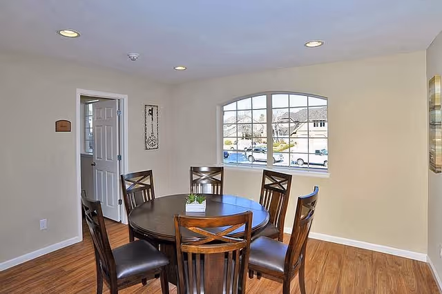 Small dining area with a round wooden table, six chairs, a potted centerpiece, recessed ceiling lights, and an arched window showing houses outside.