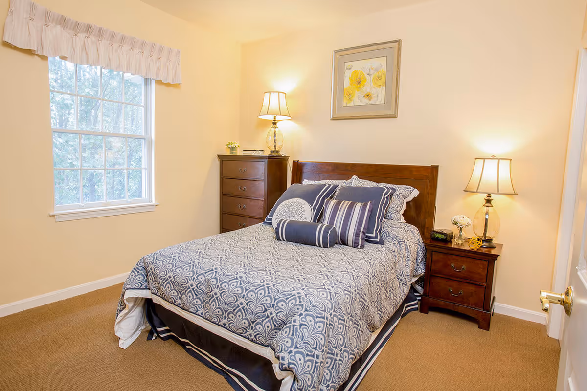 A cozy bedroom with a double bed covered in a blue and white patterned bedspread and multiple decorative pillows. There is a wooden headboard, a wooden dresser with a lamp and small plant on top, and a wooden nightstand with a lamp, clock, and decorative items. A window with a white valance lets in natural light, and a framed floral picture hangs on the wall above the bed.