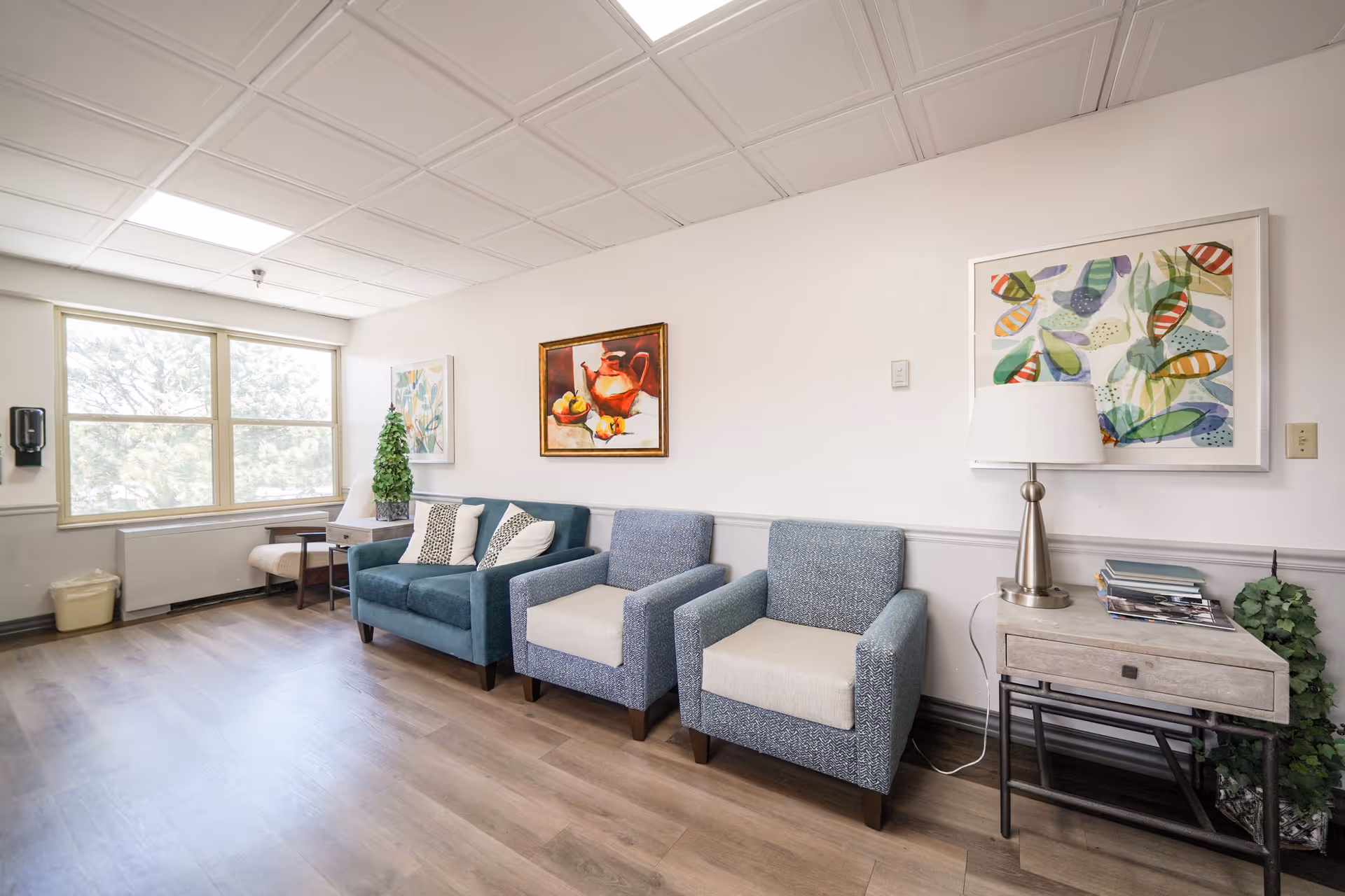 A bright and clean sitting area with three upholstered chairs, a small side table with a lamp, and two colorful paintings on the wall. Large windows allow natural light to fill the room, and there is a small potted plant on a table near the window.