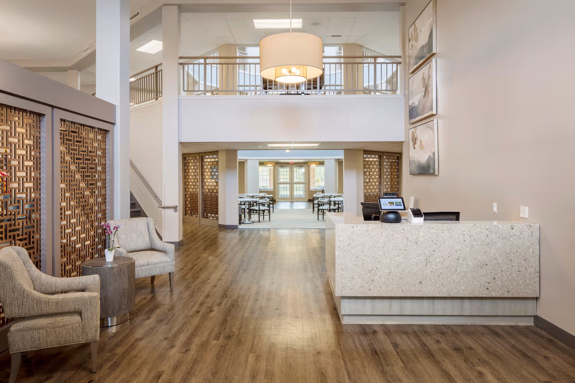 A spacious and modern reception area in a senior living facility with a light-colored stone reception desk on the right, two upholstered chairs with a small round table and a potted orchid on the left, wooden decorative panels, and a view into a dining area with tables and chairs in the background. The space features wood flooring, neutral walls, and a large hanging light fixture above.