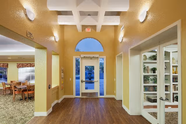 Interior view of a senior living facility hallway with wooden flooring and yellow walls. The hallway leads to a glass door with an arched window above it. On the left side, there is an open area with tables and chairs, and on the right side, there is a glass-fronted cabinet with shelves holding books and decorative items. The ceiling has a grid pattern with recessed lighting.