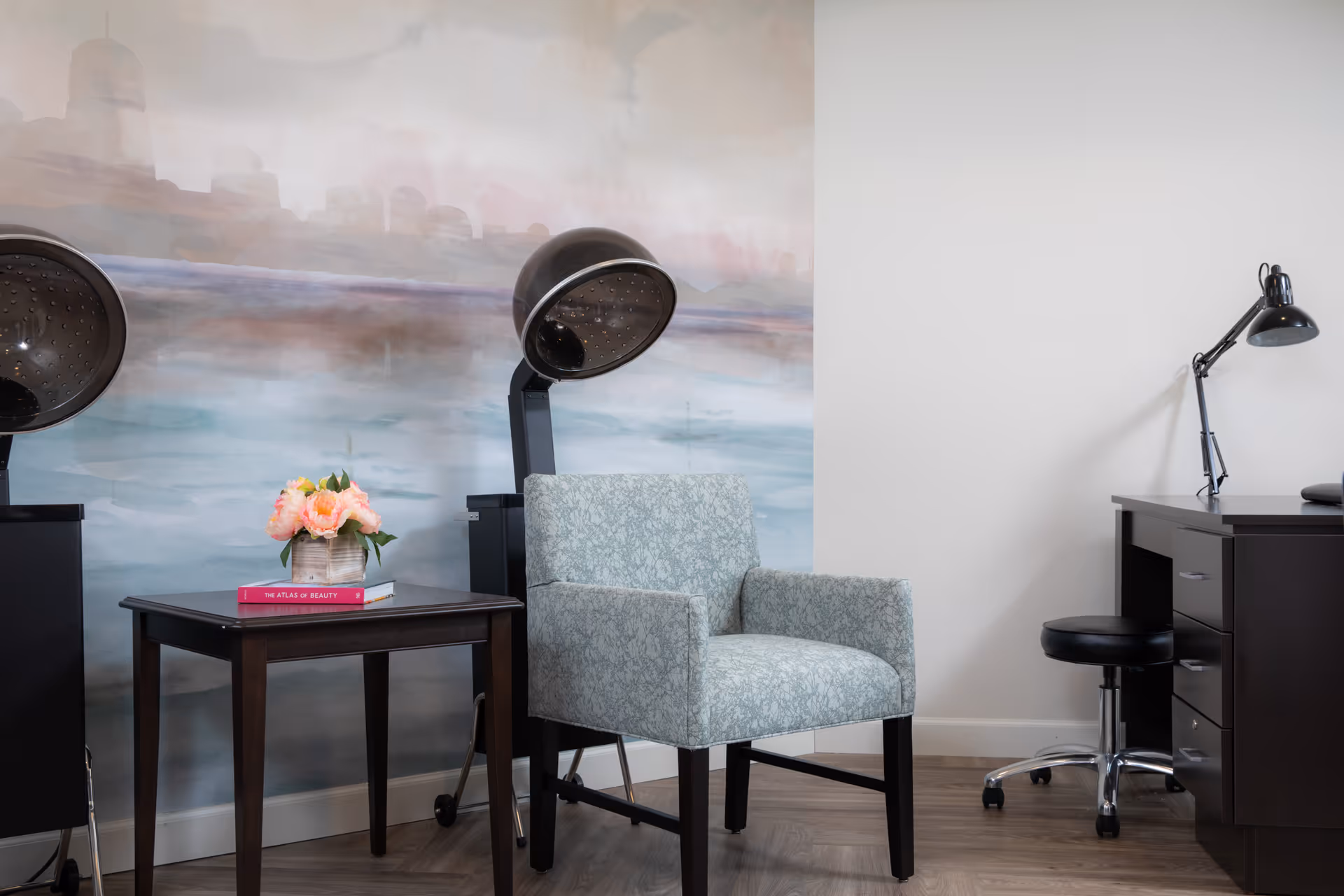 A cozy salon or beauty room with two black hair dryers on stands, a light blue patterned armchair, a small dark wooden table with a vase of pink flowers and two books, and a dark wooden desk with a black adjustable lamp and a black swivel stool. The wall behind has a soft, abstract mural in pastel colors.