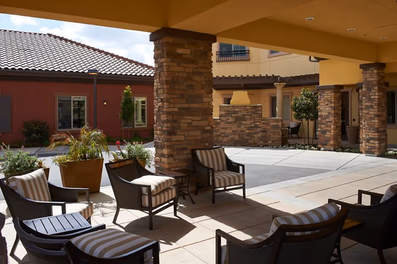 Outdoor seating area with cushioned chairs arranged around a small table under a covered patio with stone pillars. There are potted plants and a brick fireplace in the background, with buildings featuring tiled roofs and windows surrounding the courtyard.