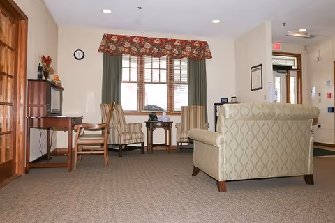 Lobby-like sitting area with upholstered armchairs, side tables, a TV cabinet and a window with a floral valance.