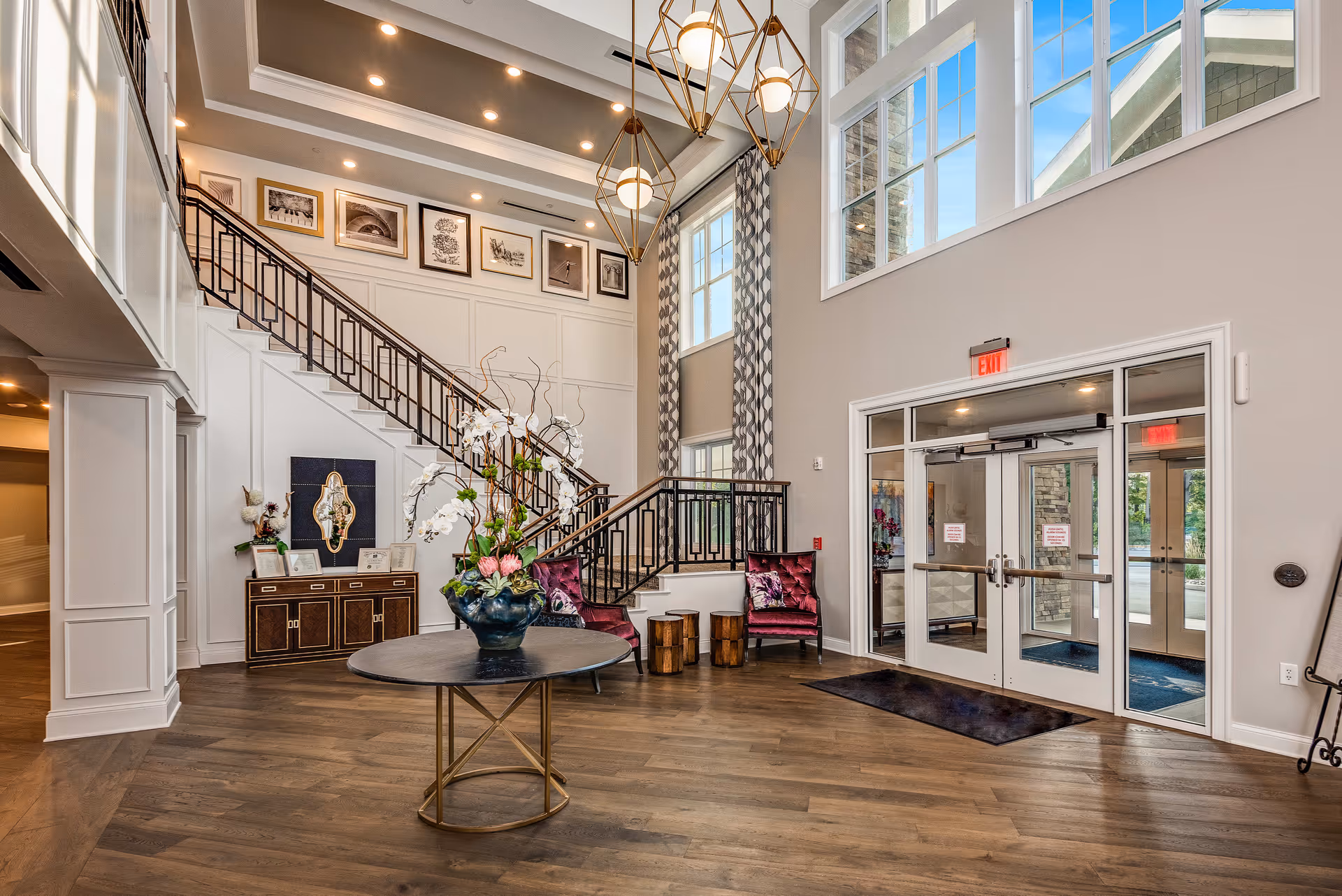 Bright two-story lobby with a decorative staircase, seating area, front glass doors, and a round table with a floral arrangement.
