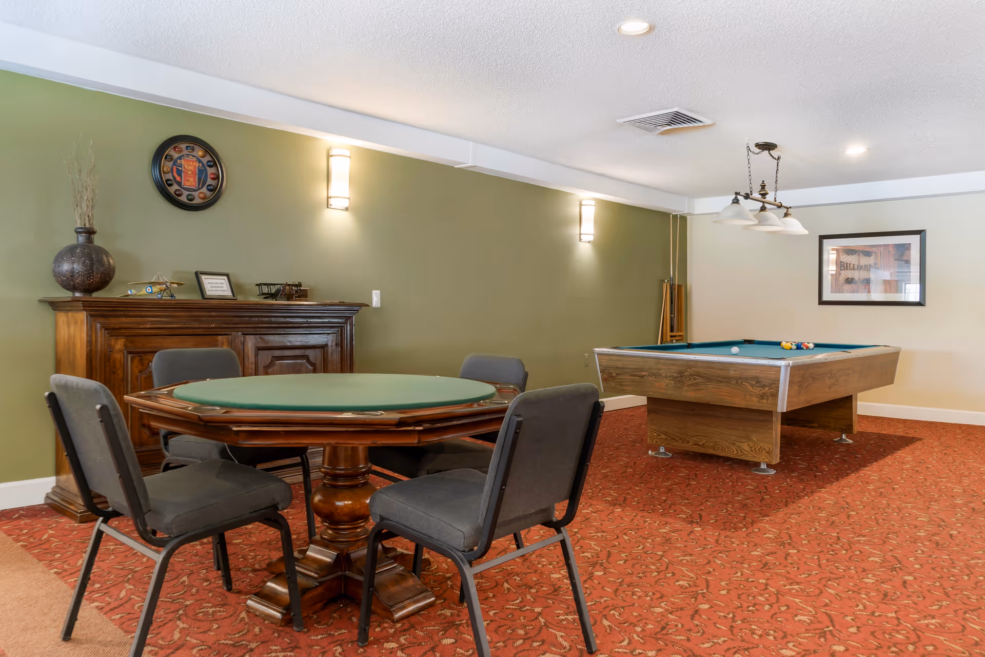 A recreational room with a green felt card table surrounded by four gray chairs in the foreground and a pool table with billiard balls and cues in the background. The room has green and beige walls, a red patterned carpet, a wooden cabinet with decorative items, and wall-mounted lights.