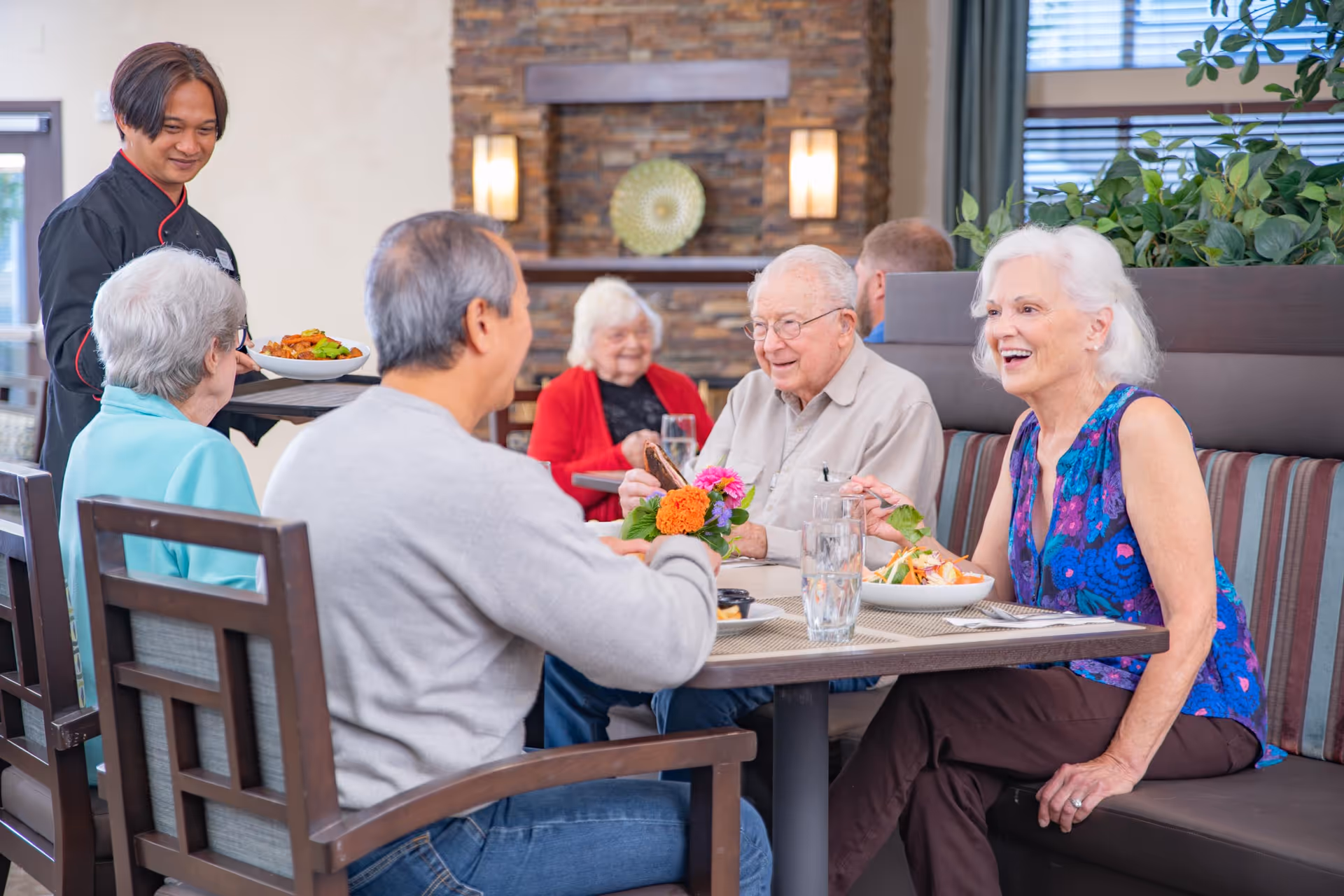 A group of elderly residents laughing and eating together at a dining table in a communal dining room while a server brings a plate.