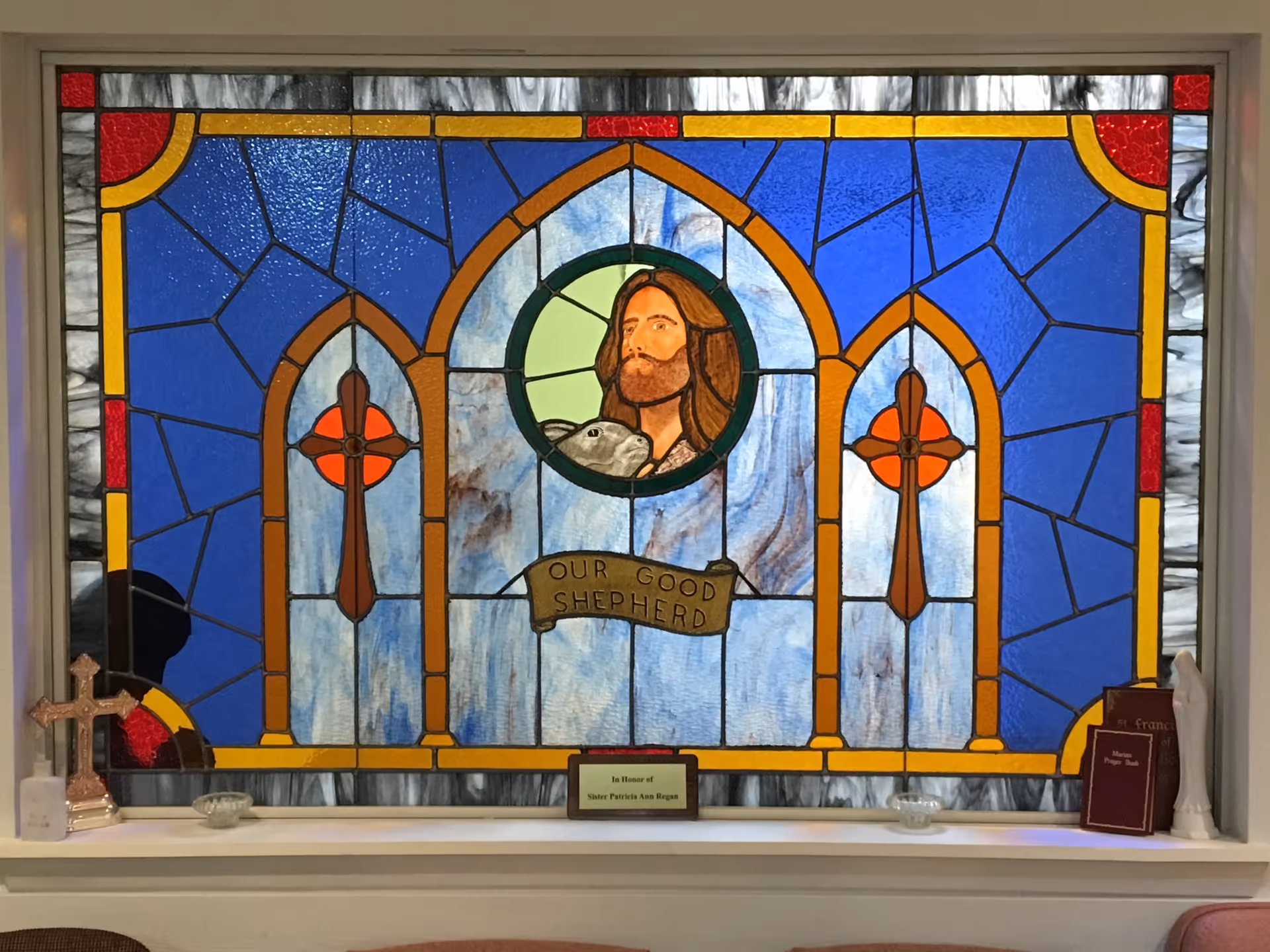 A stained glass window depicting Jesus as the Good Shepherd holding a lamb, with the words 'Our Good Shepherd' on a banner below the image. The window features blue, red, yellow, and brown colors with two crosses on either side. Below the window is a shelf with a cross, a small plaque, two prayer books, and a statue.