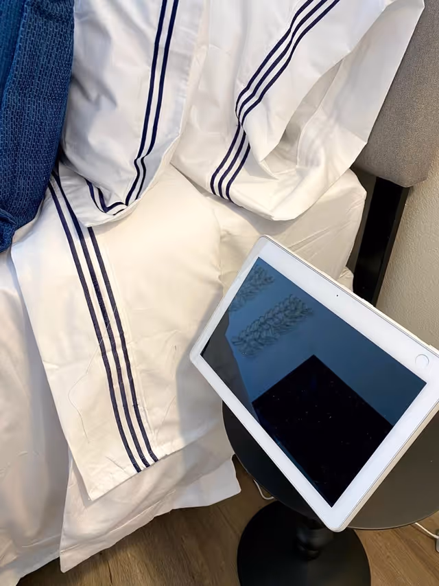 Close-up of a bed with white sheets featuring blue stripes, a blue textured pillow, and a tablet resting on a small black round side table next to the bed.