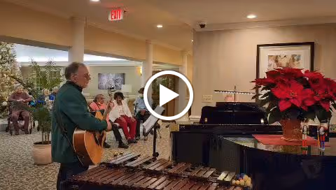 A man playing guitar and singing in a common area of a senior living facility. Several elderly residents are seated and watching the performance. The room is decorated with plants, artwork, and a poinsettia on a piano.
