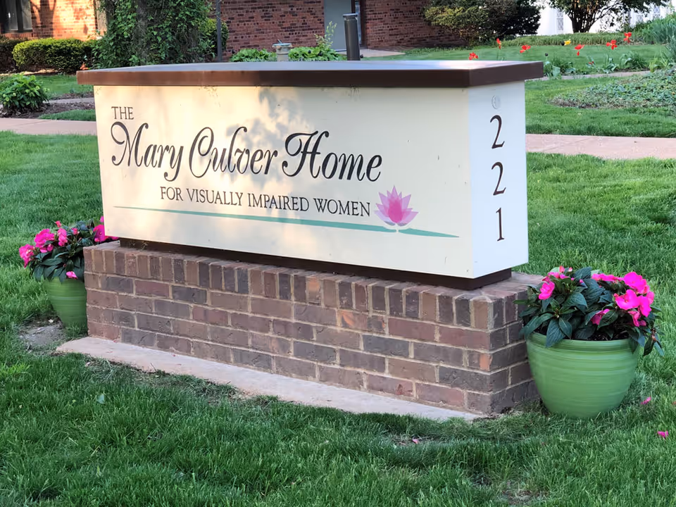 A rectangular sign mounted on a brick base on a grassy lawn. The sign reads 'The Mary Culver Home for Visually Impaired Women' with a pink lotus flower graphic and the number 221 on the side. There are two green pots with pink flowers on either side of the sign.