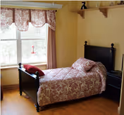 A cozy bedroom with a single dark wooden bed featuring a floral patterned bedspread and matching pillow. The room has a large window with floral curtains, a small black nightstand with a lamp, and a wooden shelf mounted on the yellow wall above the bed.