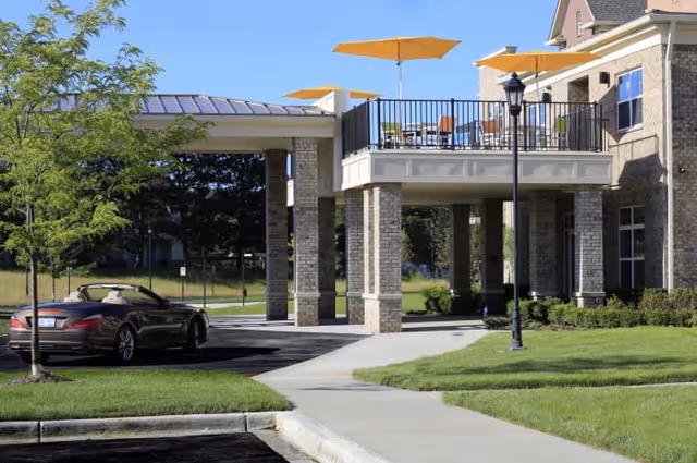 Exterior view of a senior living facility with a covered entrance supported by stone pillars. There is a balcony above the entrance with tables and chairs shaded by yellow umbrellas. A black convertible car is parked near the entrance, and there is a sidewalk and well-maintained green lawn surrounding the area.