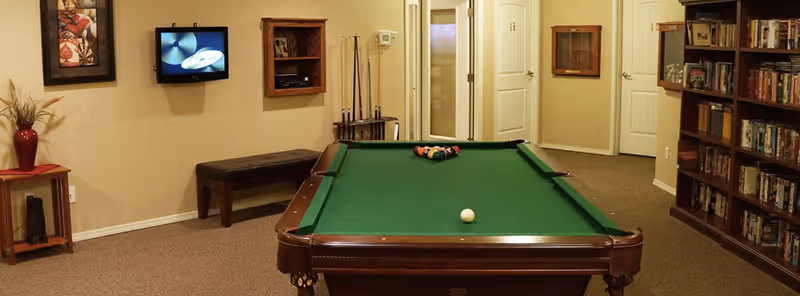 A recreation room featuring a pool table centered in the room, a wall-mounted TV, cue rack, bench, and bookshelves.