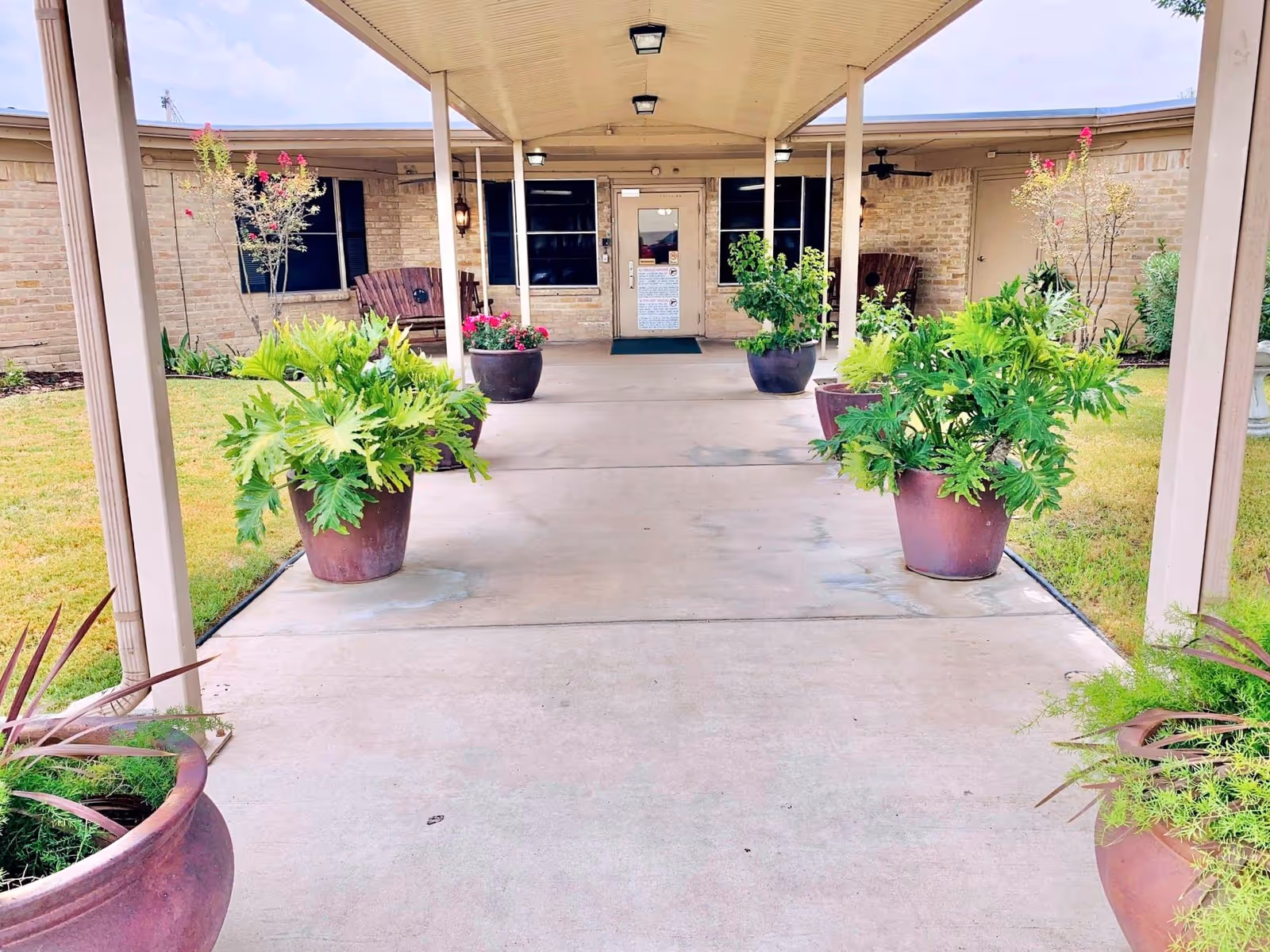 Covered entrance walkway to a single-story brick building lined with large potted plants.